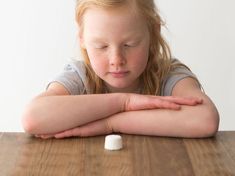 A young child leans onto a desk with arms folded and stares at a small marshmallow