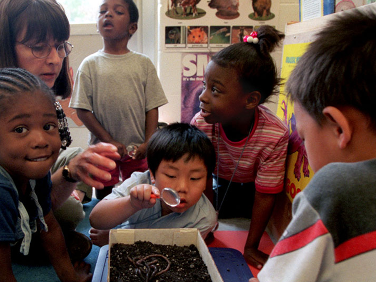 Four children are sitting around a shoe box filled with dirt and worms engaging in a science project