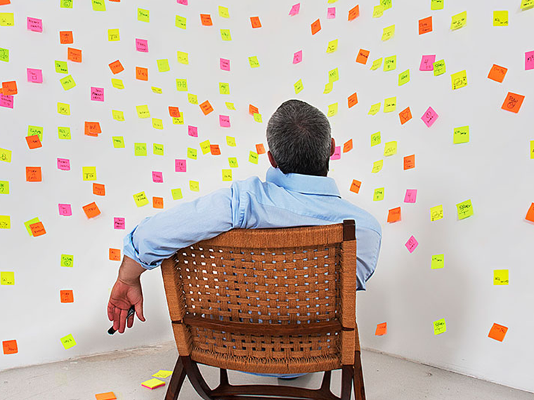 A person sits in a wicker chair looking a white wall covered in multicolored sticky notes
