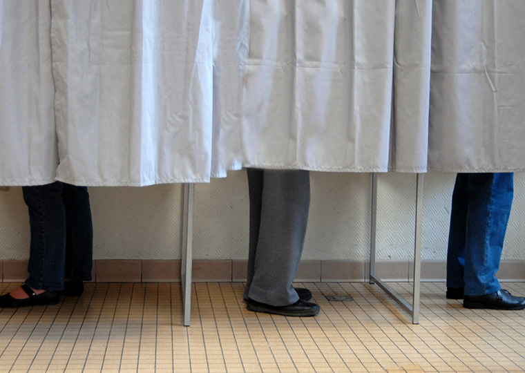 A beige curtain separates three people in a voting booth shown from below the knee to their feet