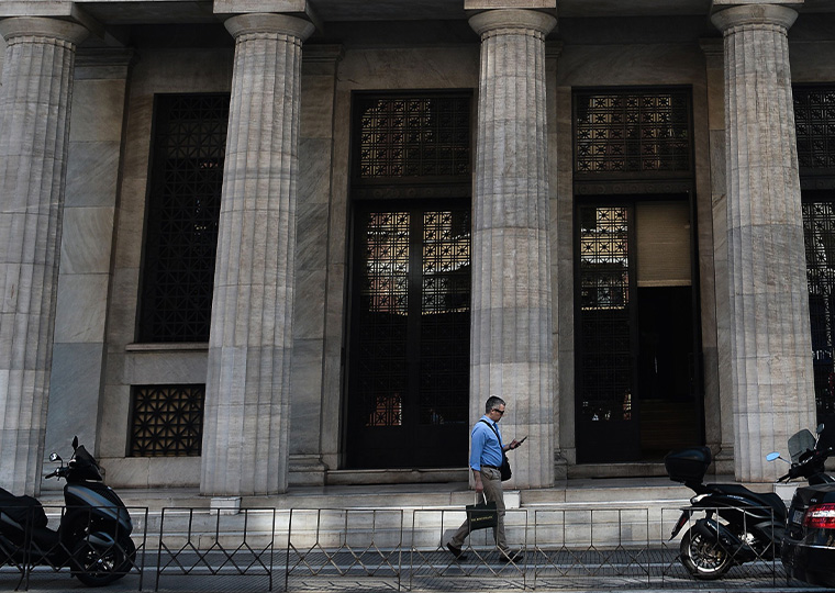 The exterior of a bank in Greece is shown as a man in a blue shirt walks in front of it.