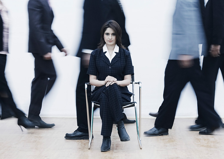 A white woman dressed in black sits in a chair while people walk behind her.