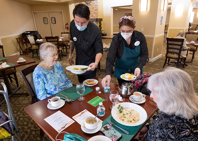 Two staffers wearing face masks hand dishes to women in the senior living center