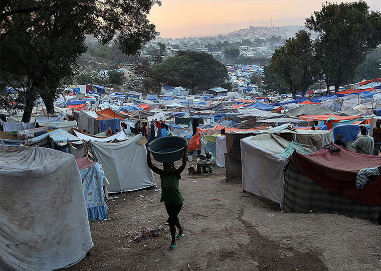 A refugee carrying a basket on their head walks through a tent encampment.