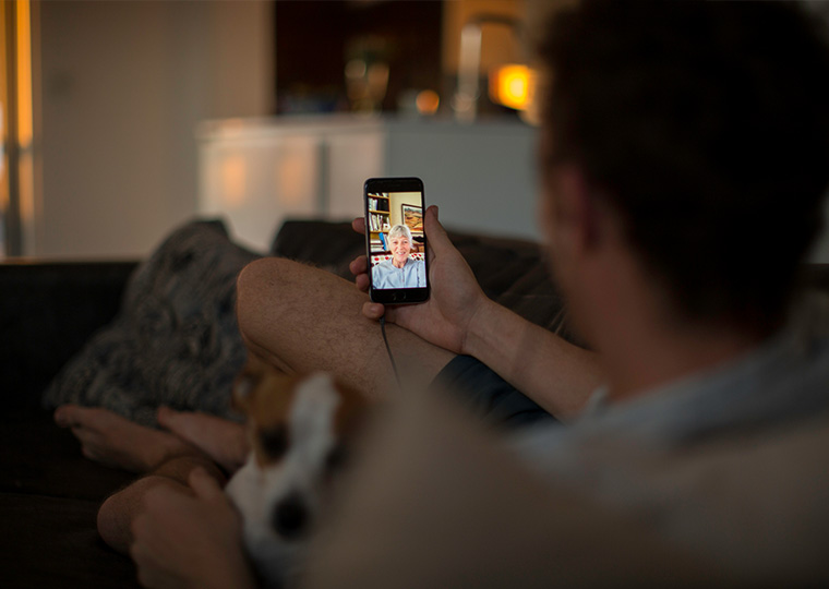 A man sitting with his dog on the couch talking to his grandma on a cellphone