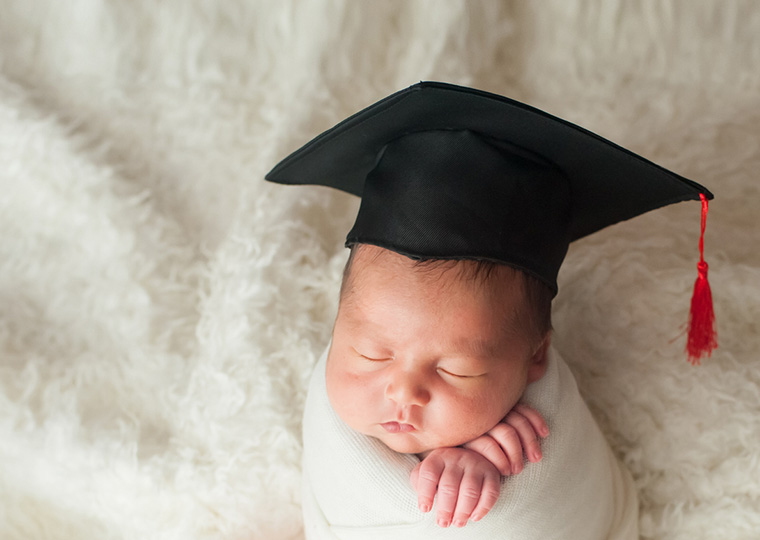 A sleeping baby wrapped in a white blanket wearing a black graduation cap with a red tasle