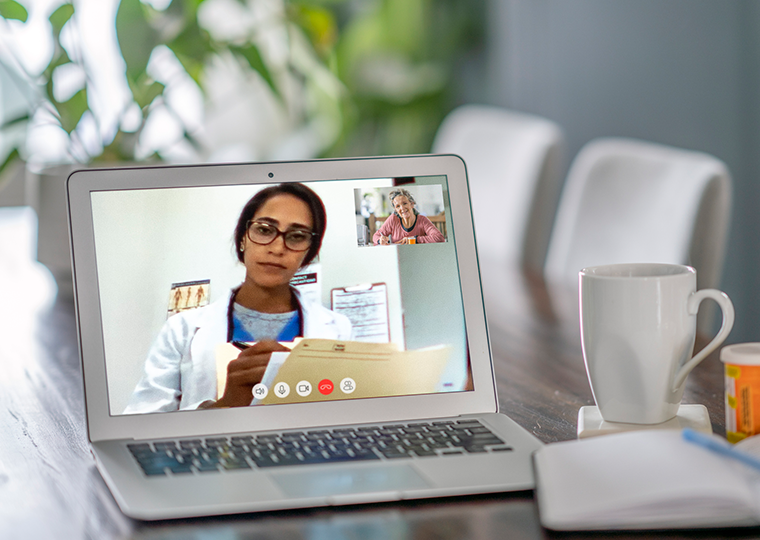 A laptop open on a table. A doctor is seen on the screen and the patient is seen in a box on the screen during a telehealth appointment.