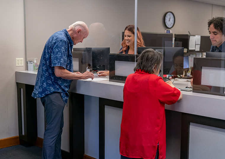 An older man fills out forms in front of a bank teller while an older woman on the right talks to a bank teller.