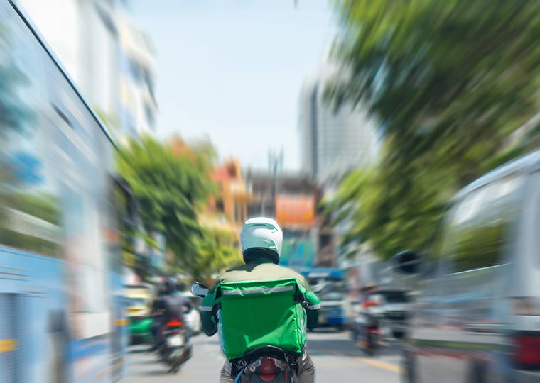 A food delivery driver in a green jacket riding a motorscooter squeezes between two vans on the street.