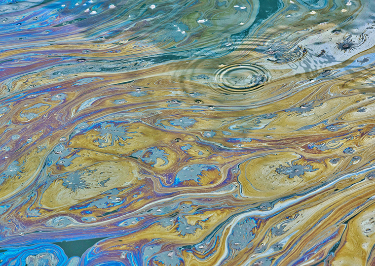 Colorful patterns on the surface of the water created by oily pollutants and stagnant water, as seen in a Texas bayou. Ripple rings also appear on the surface from aquatic life under the polluted water.