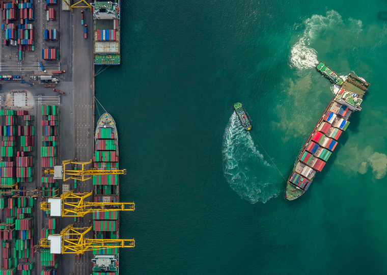 Aerial view of a cargo ship being hauled by tugboats near the port where other ships are docked and cargo waits to be loaded.