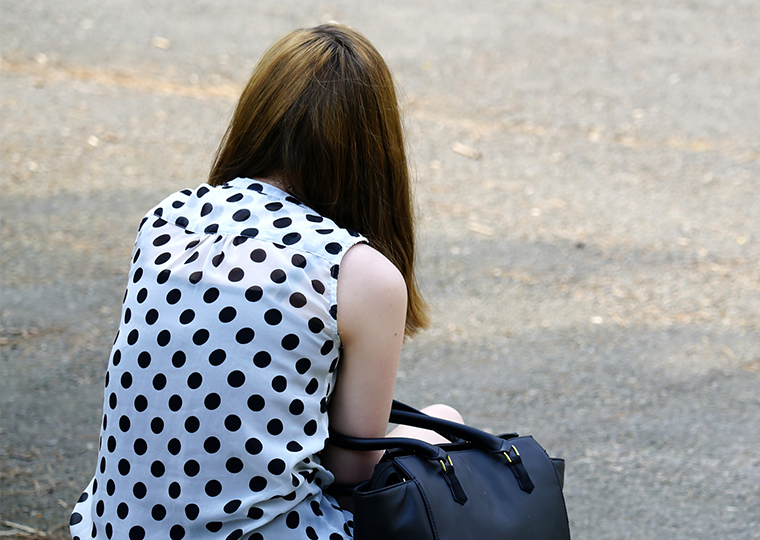 A view of the back of a young woman in white shirt with black polka dots and a black purse looped through her arm sits on stairs outside