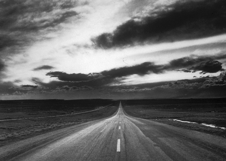 An empty two-lane highway under a cloud-filled sky at either dusk or dawn.