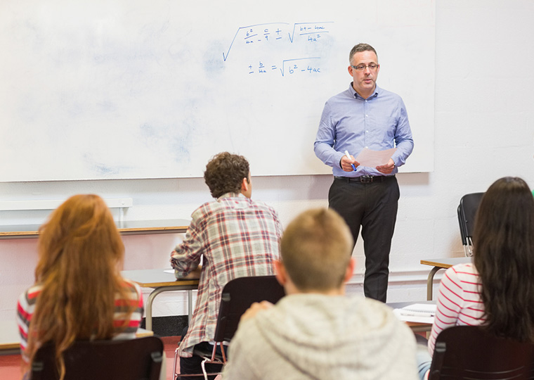 Rear view of students attentively listening to male teacher in the classroom who is standing in front of a whiteboard with a math problem written on it.