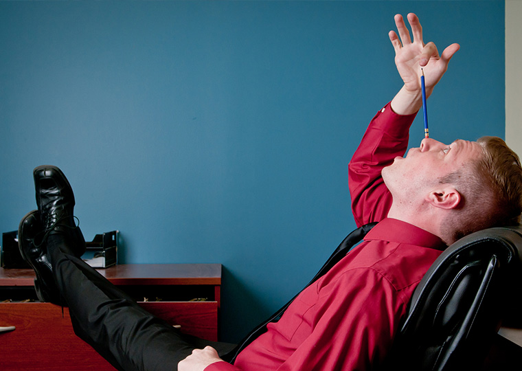 A young Caucasian male employee goofing off and being unproductive in his office when he should be working. He is in the process of trying to balance a pencil on his nose.