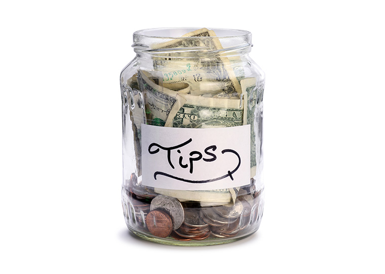A glass jar filled with change and dollar bills with a white label that reads "tips."