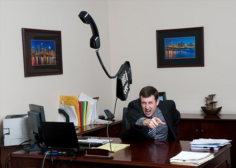 A white man wearing a business suit sitting at a desk throws his desk phone in a fit of anger.